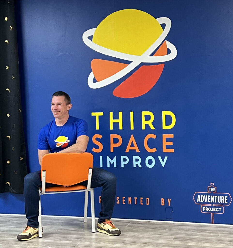 A man sits on a chair on stage wearing a Third Space Improv t-shirt.